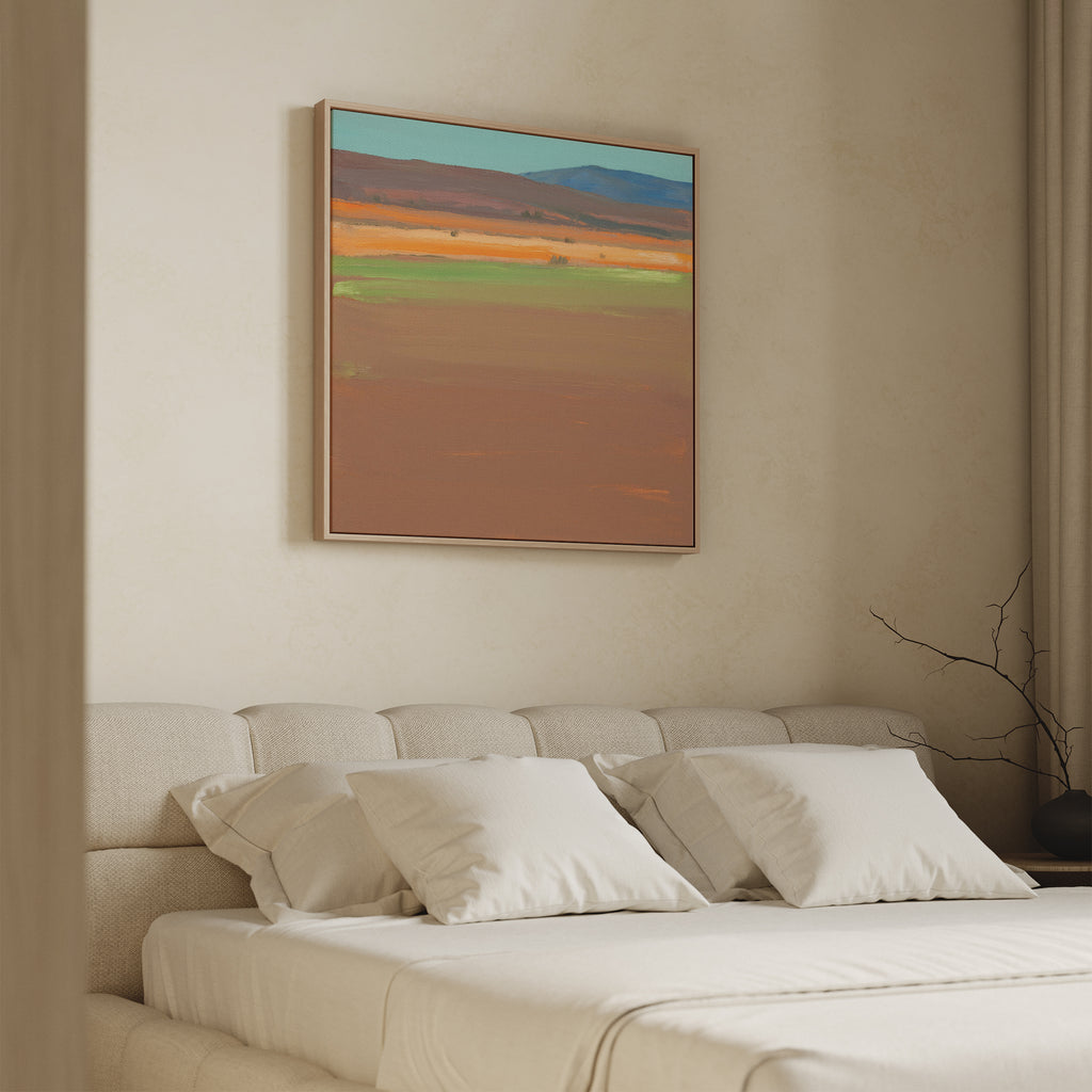 A modern bedroom features a beige upholstered bed with white bedding and four pillows. Above the bed hangs the Desert Plains Canvas Art - Square. A minimalist vase with twigs on the bedside table enhances the calm atmosphere.