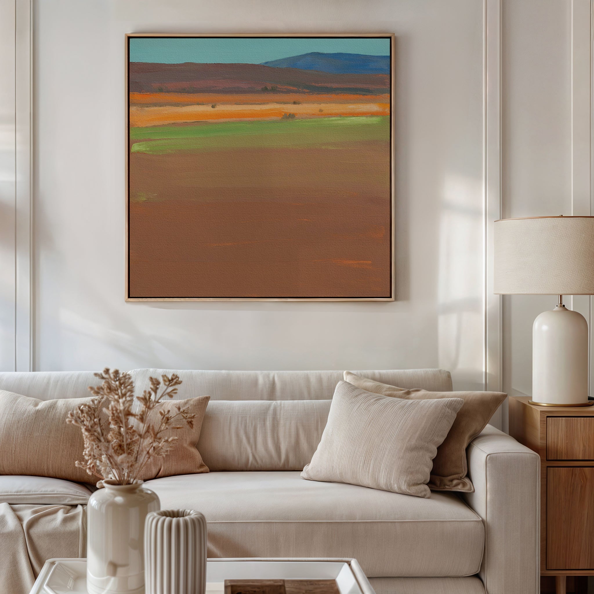 A modern living room with a beige sofa, neutral cushions, a lamp on a wooden side table, vases with dried plants, and the Desert Plains Canvas Art - Square—a colorful landscape—hanging on the white wall above the couch.