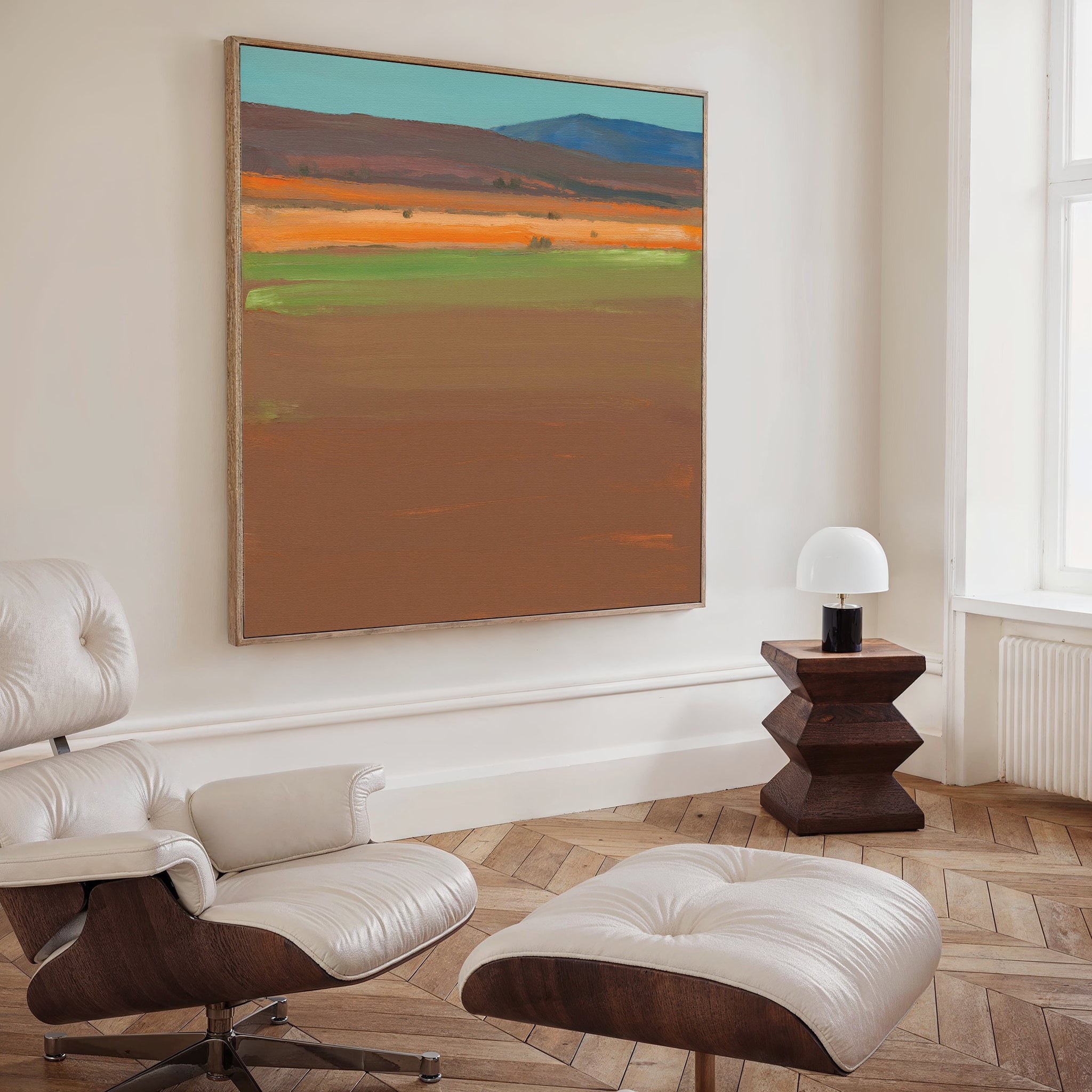 A modern living room with a white lounge chair, matching ottoman, dark wood side table with a white lamp, and the Desert Plains Canvas Art - Square featuring earthy tones and blue accents. Perfect modern living room decor.