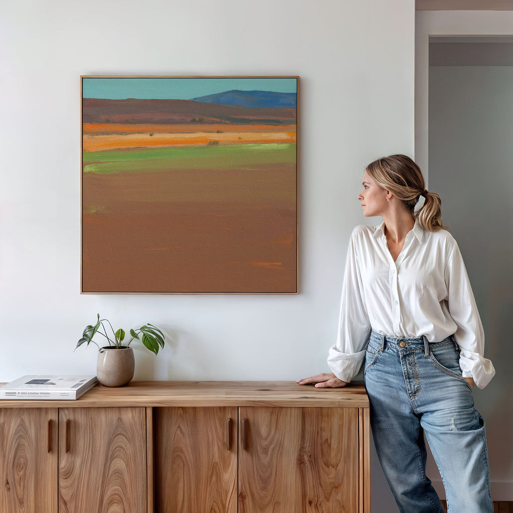 A woman in a white blouse and blue jeans stands by a wooden cabinet, admiring the Desert Plains Canvas Art - Square with earthy southwestern tones. A potted plant and books add charm to the modern living room decor.