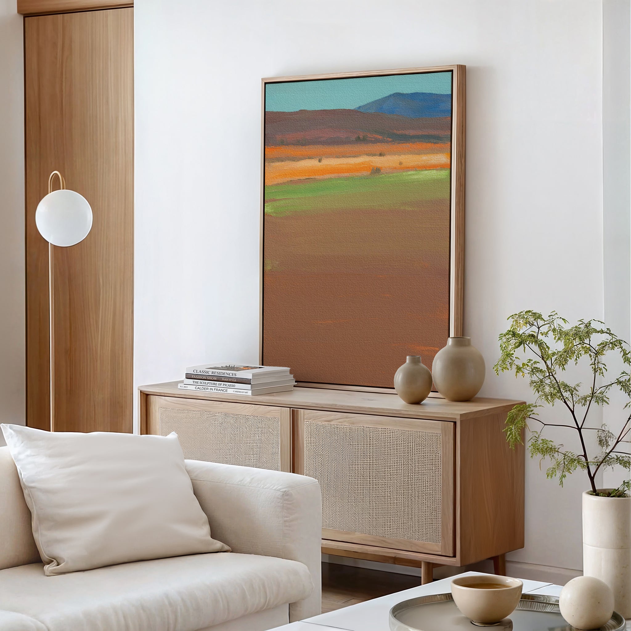A modern living room features a beige sofa, light wood sideboard, pottery vases, stacked books, a small potted plant, round pendant lamp, and Desert Plains Canvas Art - Vertical—a striking southwestern wall art accent for the space.