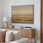 A cozy living room corner features a light wood sideboard with books, a lamp, and decor. Above the beige sofa with white cushions, the "Desert Solitude Canvas Art - Horizontal" hangs on the white wall as serene wall decor.