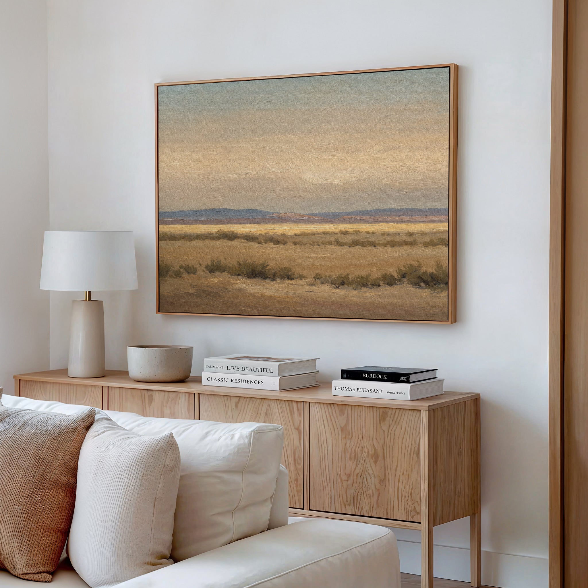 A cozy living room corner features a light wood sideboard with books, a lamp, and decor. Above the beige sofa with white cushions, the "Desert Solitude Canvas Art - Horizontal" hangs on the white wall as serene wall decor.