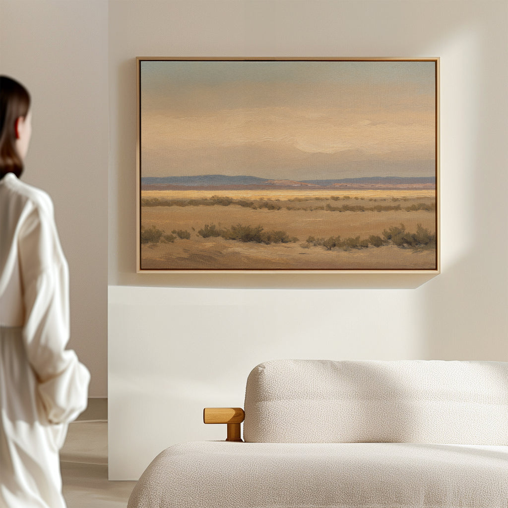 A person in a white shirt stands in a bright, minimal room, gazing at the Desert Solitude Canvas Art - Horizontal—a southwestern landscape print with calm desert and distant mountains—hanging above a simple cream-colored sofa.