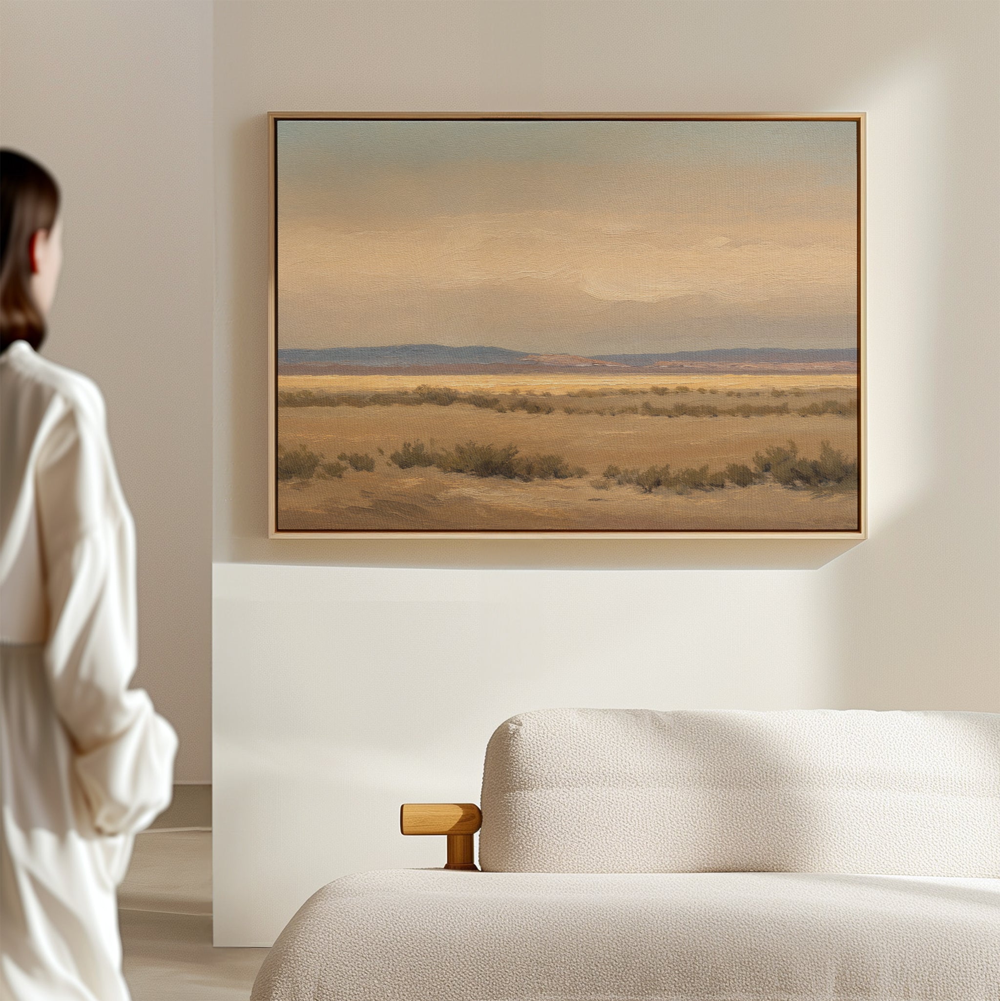 A person in a white shirt stands in a bright, minimal room, gazing at the Desert Solitude Canvas Art - Horizontal—a southwestern landscape print with calm desert and distant mountains—hanging above a simple cream-colored sofa.