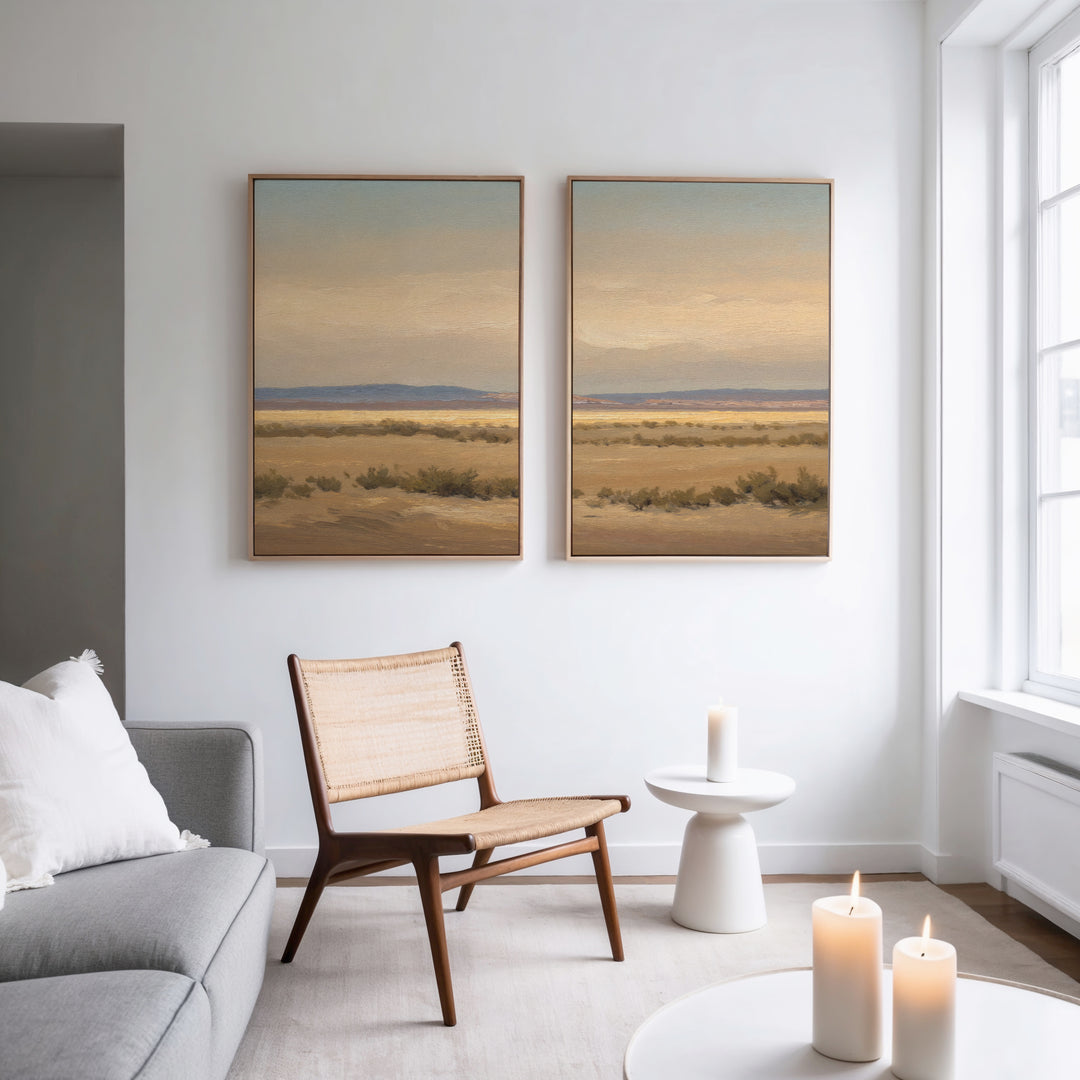 A bright, minimalist living room with a gray sofa, woven chair, and small round table with a lit candle. Serene southwestern landscape art, Desert Solitude - Set of 2, hangs above. More candles glow on a foreground table.