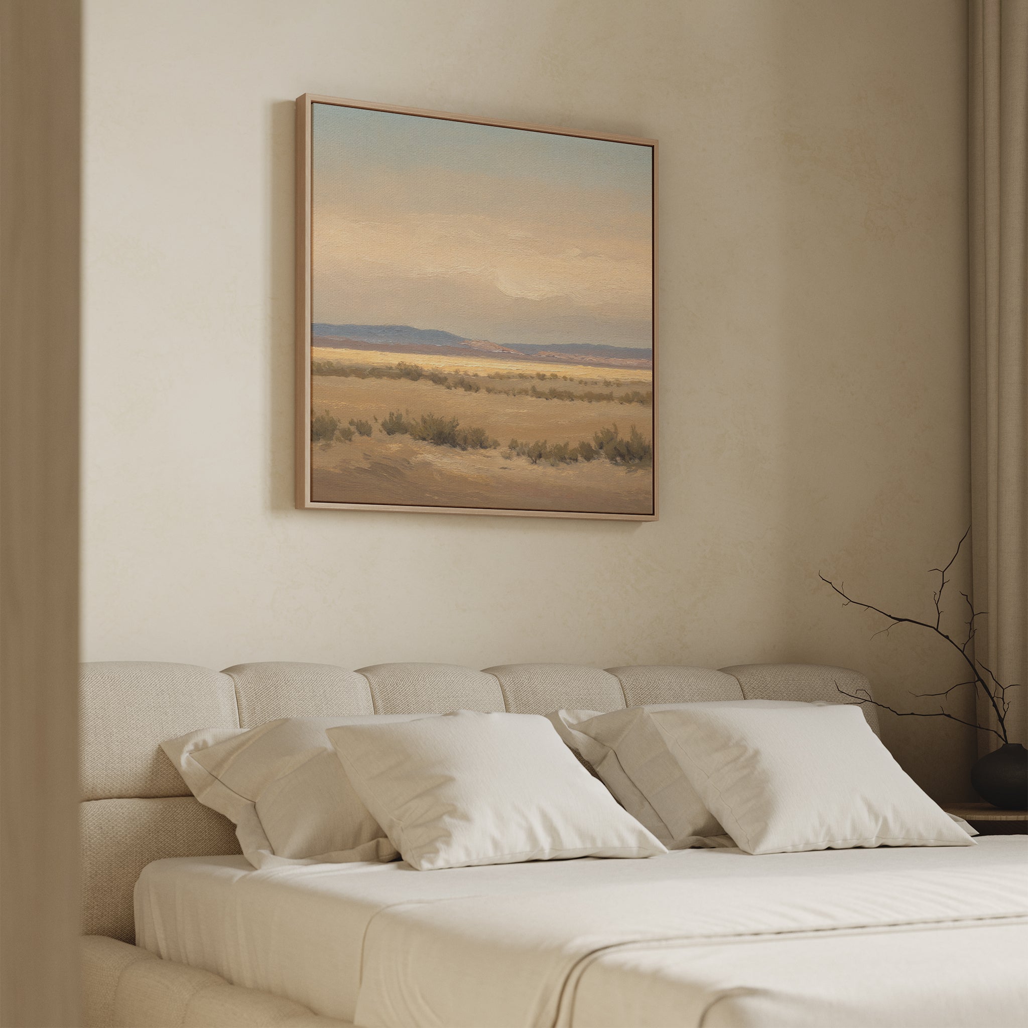A neatly made bed with white bedding sits against a beige wall. Above it hangs the Desert Solitude Canvas Art - Square, featuring a southwestern landscape with distant mountains. A bare branch decorates the bedside table.