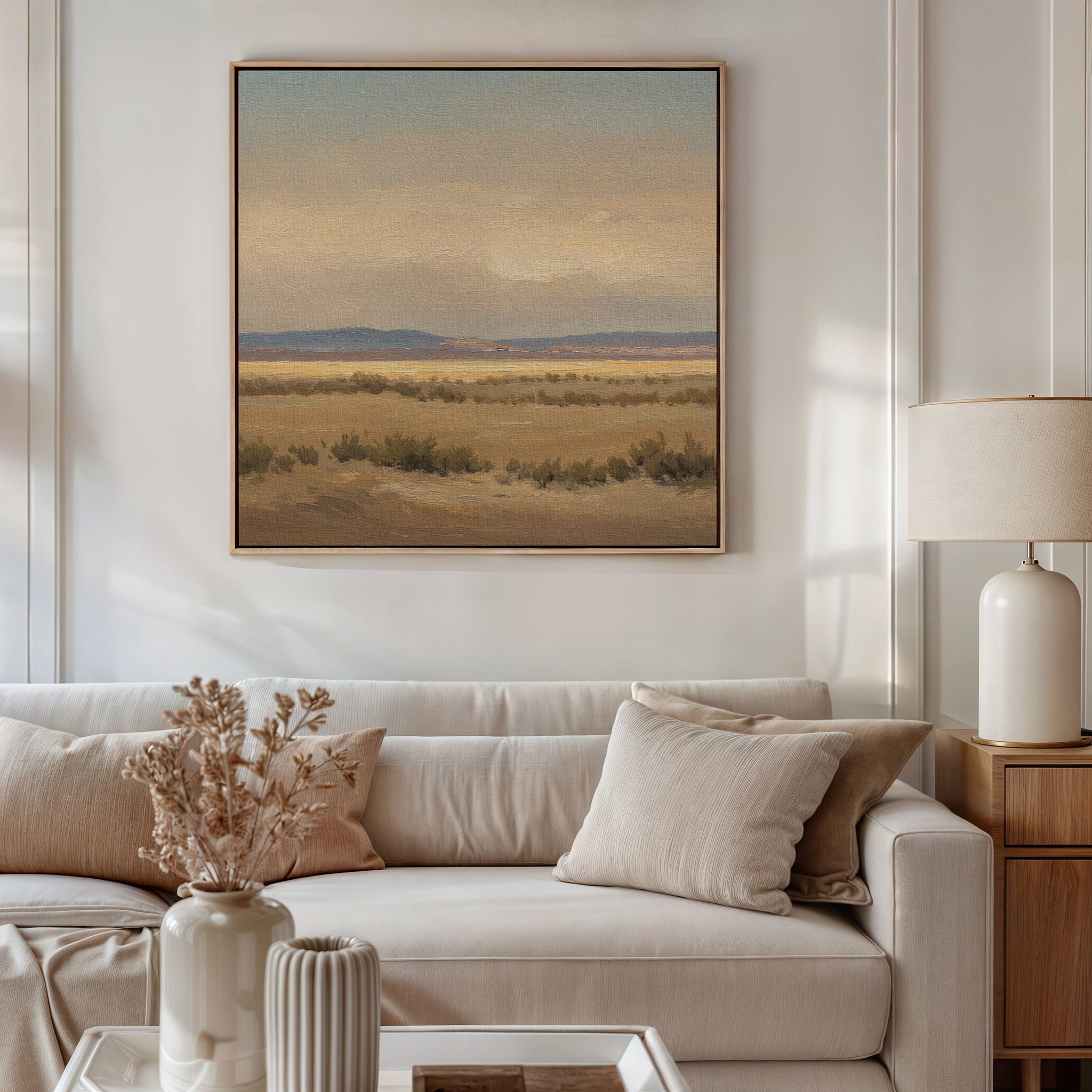 A beige sofa with neutral cushions sits in a bright living room. Above, the Desert Solitude Canvas Art - Square adds serene charm, while a table lamp, wooden side table, and dried flowers in a vase complete the cozy decor.