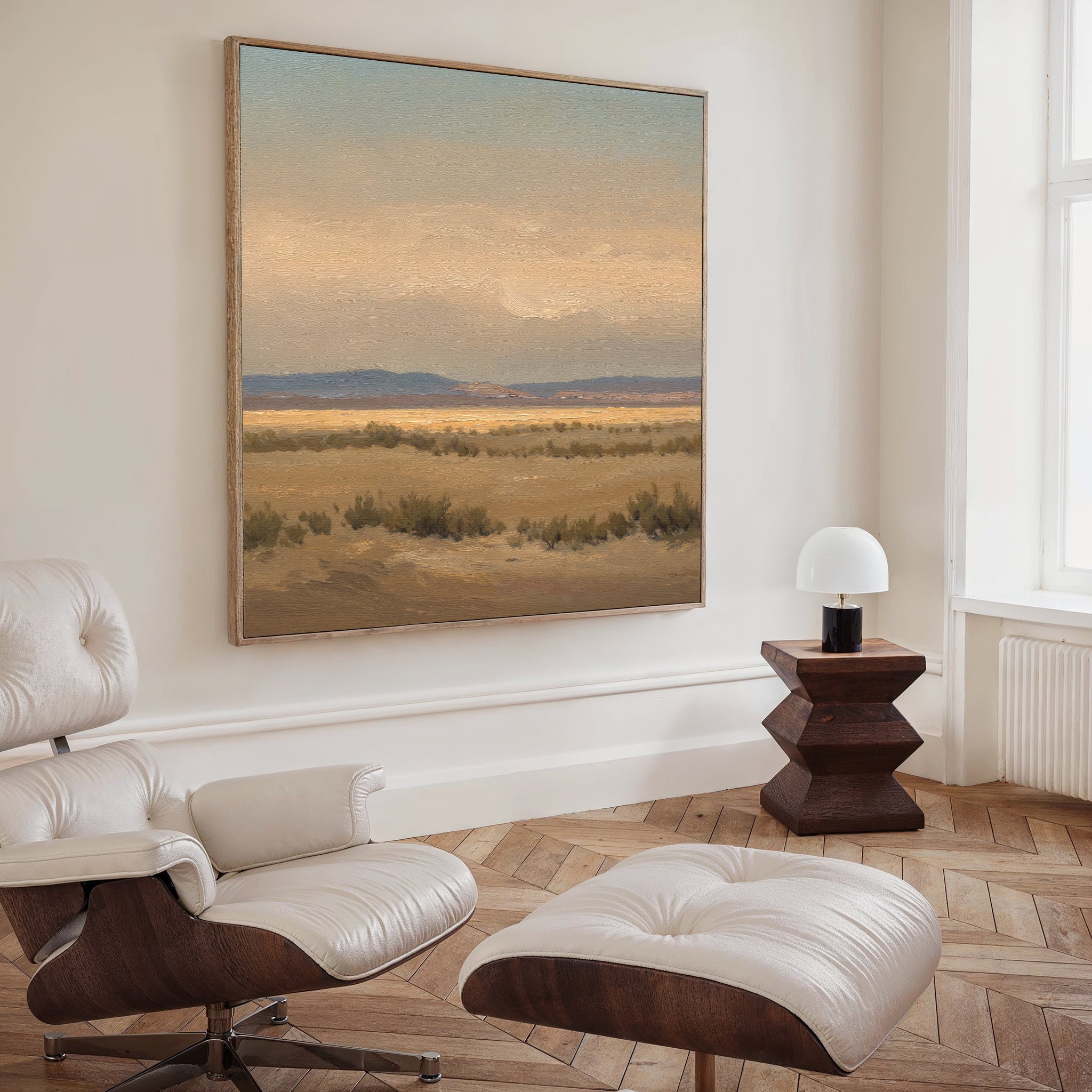 A modern living room featuring a white leather lounge chair and ottoman, a geometric wooden side table with a small lamp, and serene wall decor with the Desert Solitude Canvas Art - Square depicting a field and mountains.