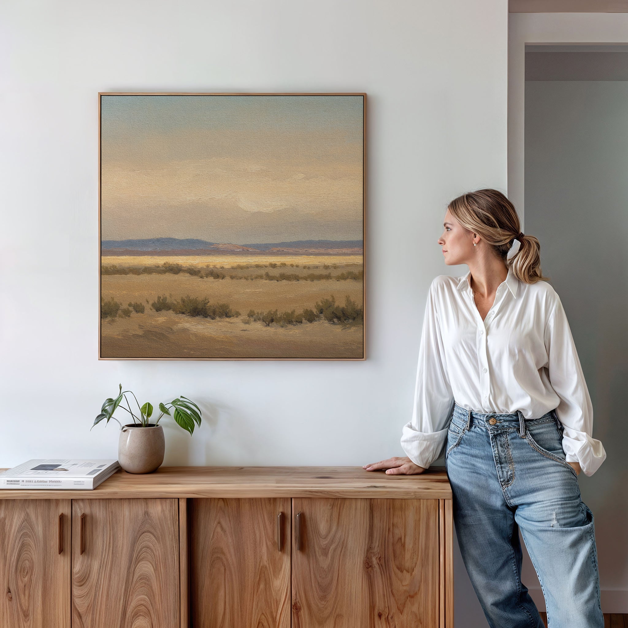 A woman in a white blouse and jeans stands by a wooden cabinet, admiring the Desert Solitude Canvas Art - Square, featuring a serene southwestern landscape scene, while a plant and books sit on the cabinet.