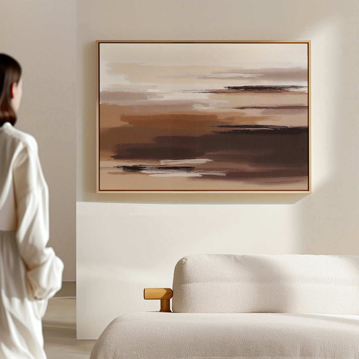 A person stands in a minimal, light-filled room with a modern beige sofa and the Desert Winds Canvas Art - Horizontal, an abstract earth-toned print in warm brown and cream, hanging on the wall.