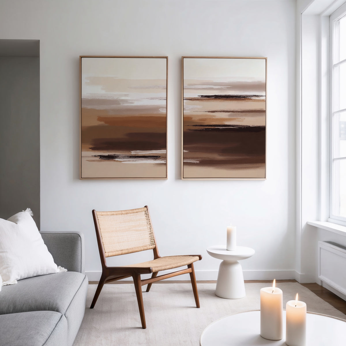 A minimalist living room with a woven chair, a round side table holding a lit candle, and the Desert Winds - Set Of 2 abstract prints in brown tones on the wall; natural light pours in from tall windows.