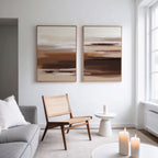 A minimalist living room with a woven chair, a round side table holding a lit candle, and the Desert Winds - Set Of 2 abstract prints in brown tones on the wall; natural light pours in from tall windows.