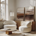 A modern living room featuring the Desert Winds - Set Of 2 white curved armchairs, a round wooden coffee table, and earth-toned artwork on a wooden sideboard. Large windows bathe the space in natural light.