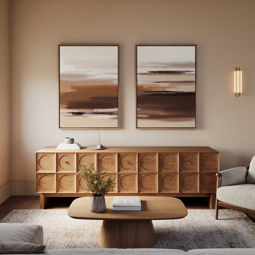 A modern living room features the Desert Winds - Set Of 2 earth-toned artworks in beige and brown, a wooden sideboard, round coffee table with book and vase, light armchair, and a wall sconce on a neutral wall.