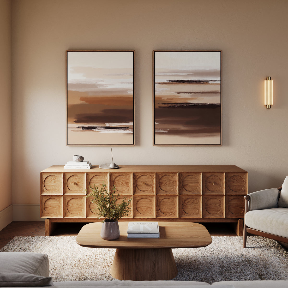 A modern living room features the Desert Winds - Set Of 2 earth-toned artworks in beige and brown, a wooden sideboard, round coffee table with book and vase, light armchair, and a wall sconce on a neutral wall.