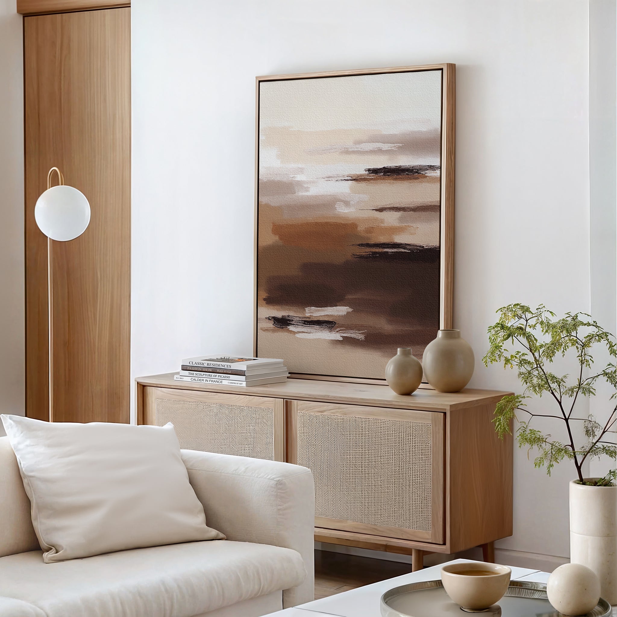 A minimalist living room with a light wooden sideboard, Desert Winds Canvas Art - Vertical, neutral vases, stacked books, a white sofa with a cushion, a small potted plant, and a round hanging light—perfect for Nordic Park style.
