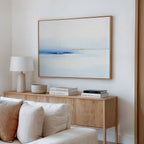 A light, modern living room featuring a wooden sideboard, white lamp, stacked books, a bowl, and the Distant Archipelago Canvas Art - Horizontal in soft blue and beige tones hanging on the white wall above.