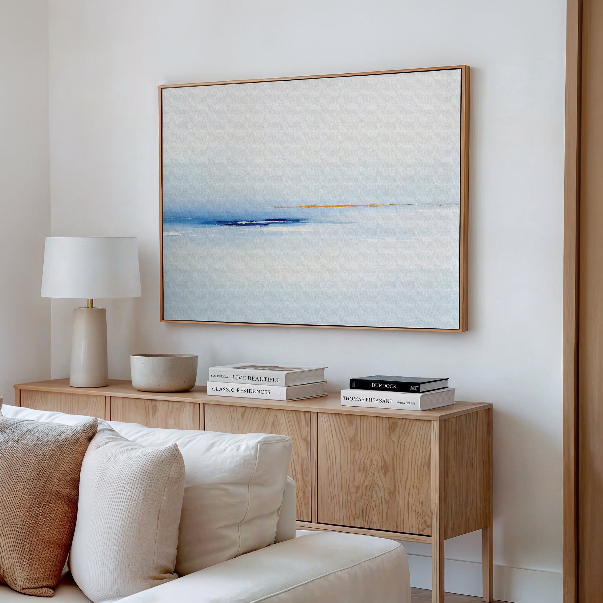 A light, modern living room featuring a wooden sideboard, white lamp, stacked books, a bowl, and the Distant Archipelago Canvas Art - Horizontal in soft blue and beige tones hanging on the white wall above.