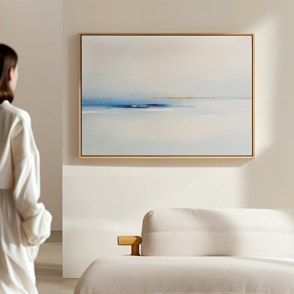 A person in a white outfit stands in a minimalist room, admiring the Distant Archipelago Canvas Art - Horizontal with soft blue and white tones hanging above a beige sofa, enhancing the elegant feel of the light-colored space.