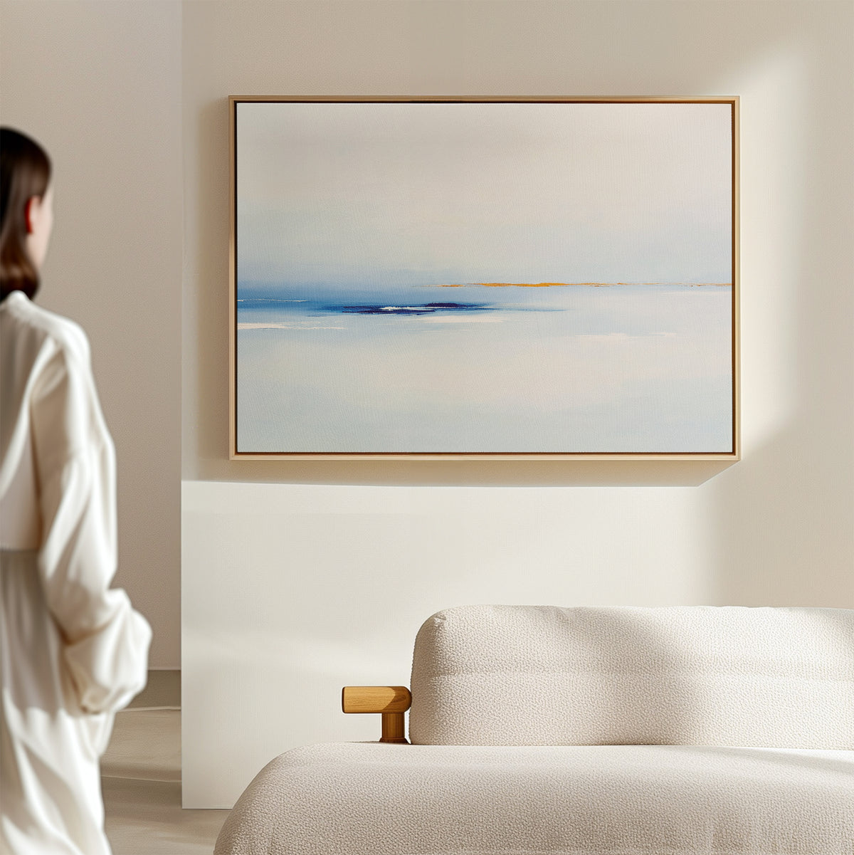 A person in a white outfit stands in a minimalist room, admiring the Distant Archipelago Canvas Art - Horizontal with soft blue and white tones hanging above a beige sofa, enhancing the elegant feel of the light-colored space.