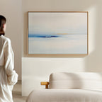 A person in a white outfit stands in a minimalist room, admiring the Distant Archipelago Canvas Art - Horizontal with soft blue and white tones hanging above a beige sofa, enhancing the elegant feel of the light-colored space.