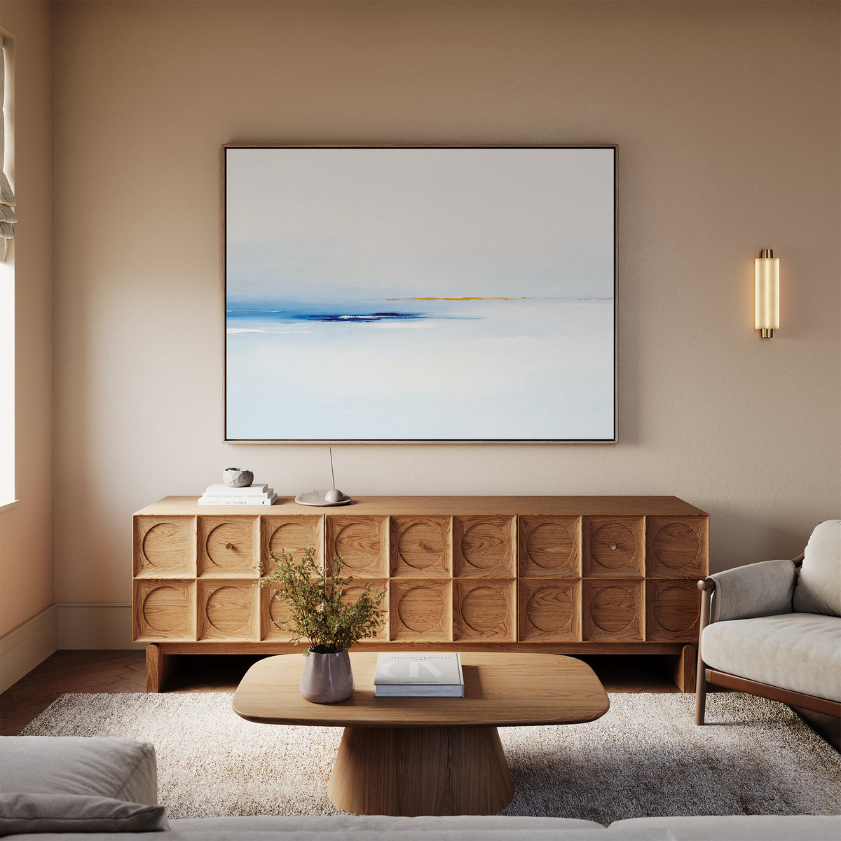 A modern living room with a light wood coffee table, beige sofa, textured wood sideboard, and Distant Archipelago Canvas Art - Horizontal in blue and white tones, plus a wall sconce, set against neutral walls and carpet.