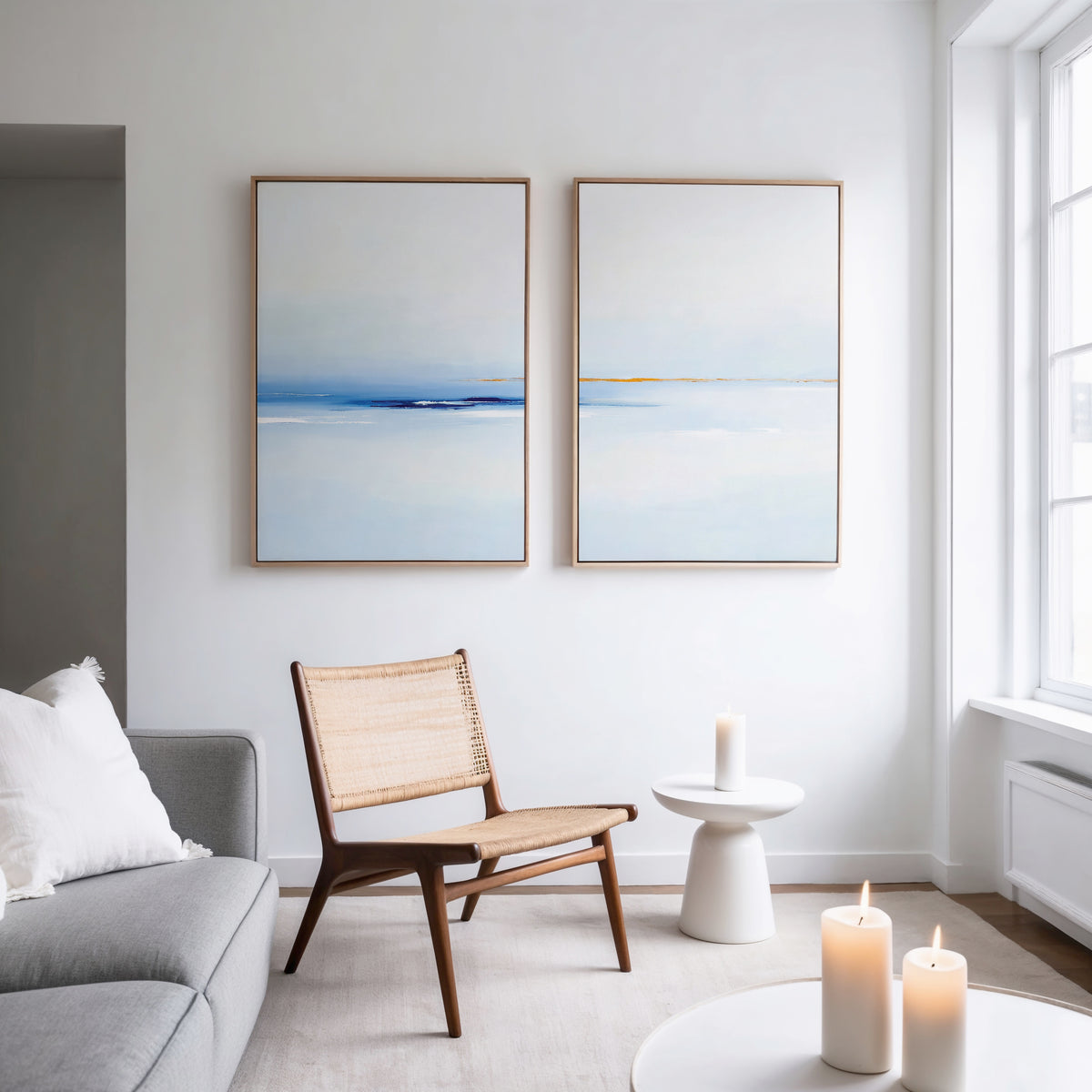 A modern, minimal living room with a wooden chair, gray sofa, small white side table with a lit candle, and "Distant Archipelago - Set Of 2" blue and white wall art above the sofa. Three candles sit on a low table near a large window.
