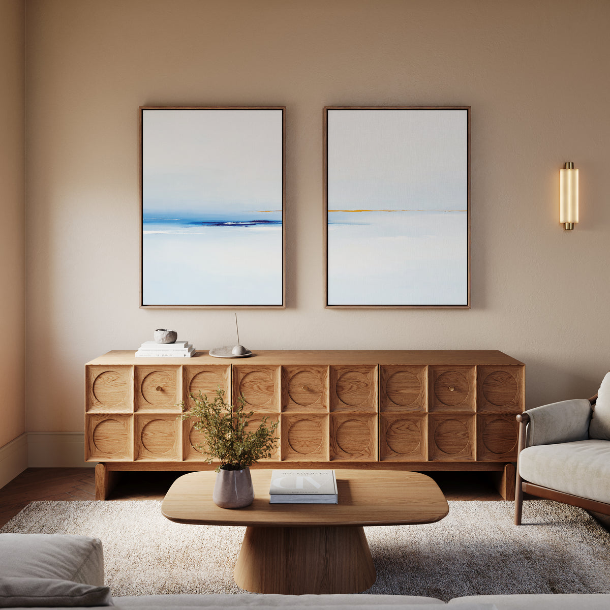 A modern living room featuring a wooden sideboard, the Distant Archipelago - Set Of 2 wall art above it, a round wooden coffee table with a book and vase, and a light gray armchair, all in neutral tones.