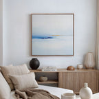 A modern, minimalist living room features a beige sofa, neutral pillows, wooden sideboard, decorative vases, stacked books, and the Distant Archipelago Canvas Art - Square in striking blue and white tones displayed on the wall.