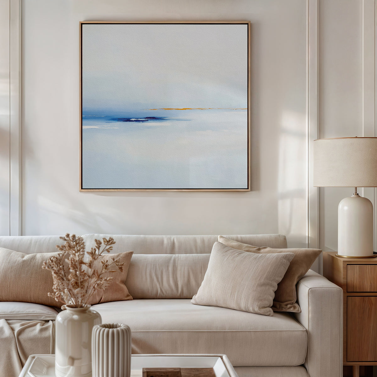 A modern living room features a beige sofa, cream cushions, a wooden side table with a lamp, dried flowers in a vase, and the Distant Archipelago Canvas Art - Square displaying soft blue and white tones on the wall.