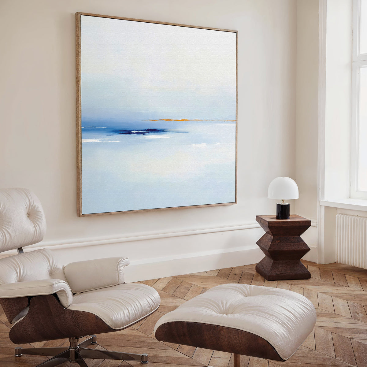 A modern living room with a white leather lounge chair and ottoman, wooden floors, a geometric side table with a lamp, and contemporary wall art—Distant Archipelago Canvas Art - Square—displayed on the wall.