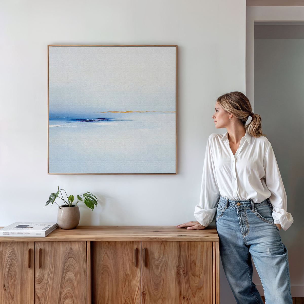 A woman in a white blouse and jeans leans against a wooden sideboard, gazing at the Distant Archipelago Canvas Art - Square hanging above her. A small plant and books add charm to the serene, modern setting.