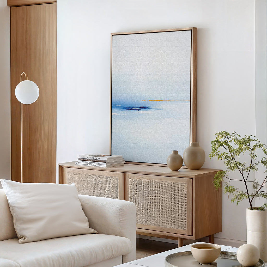 Minimalist living room featuring a light wood sideboard, Distant Archipelago Canvas Art - Vertical in blue and white, neutral vases, stacked books, a white sofa with pillows, a globe floor lamp, and a small potted plant.