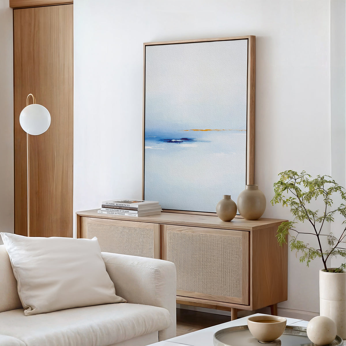Minimalist living room featuring a light wood sideboard, Distant Archipelago Canvas Art - Vertical in blue and white, neutral vases, stacked books, a white sofa with pillows, a globe floor lamp, and a small potted plant.