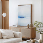 Minimalist living room featuring a light wood sideboard, Distant Archipelago Canvas Art - Vertical in blue and white, neutral vases, stacked books, a white sofa with pillows, a globe floor lamp, and a small potted plant.