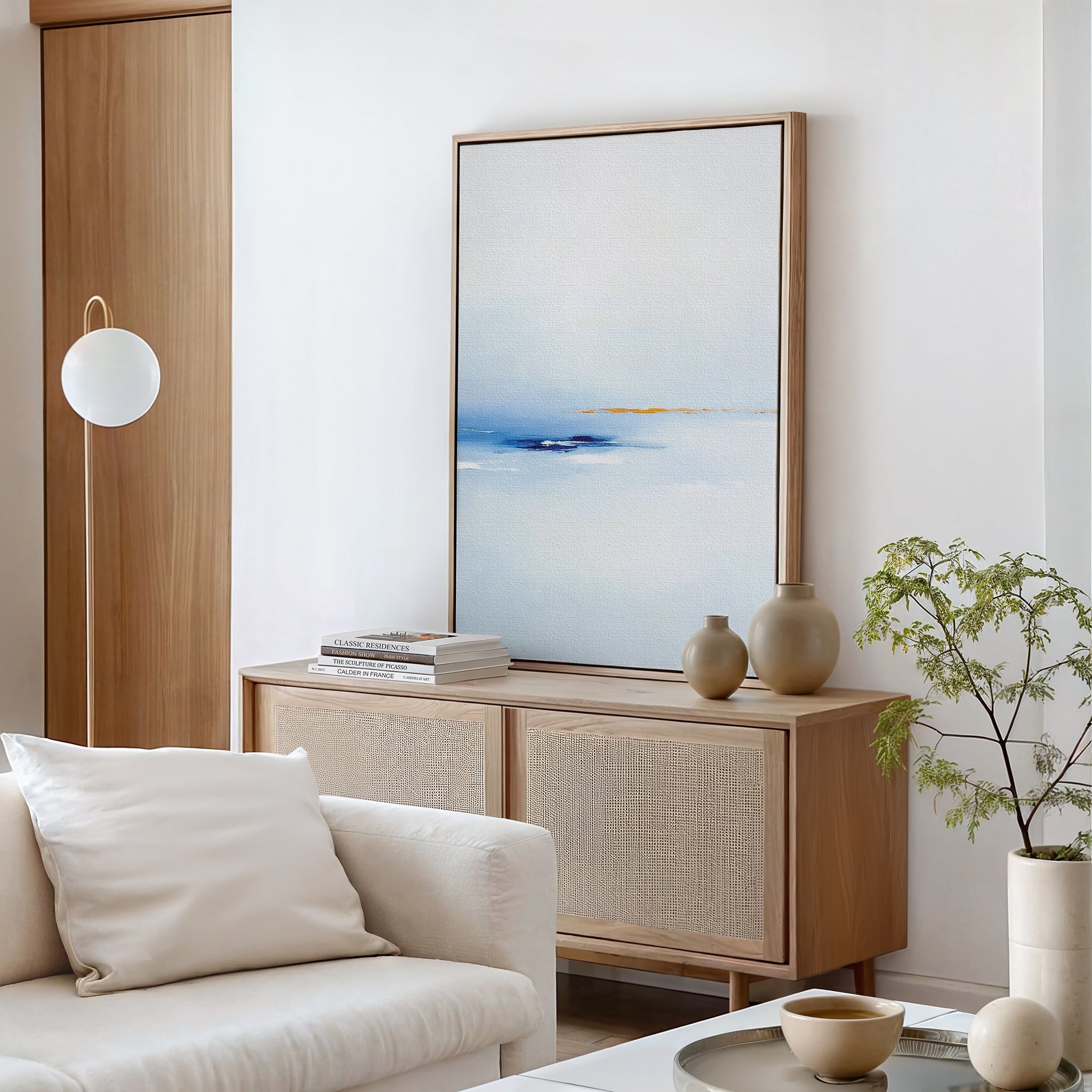 Minimalist living room featuring a light wood sideboard, Distant Archipelago Canvas Art - Vertical in blue and white, neutral vases, stacked books, a white sofa with pillows, a globe floor lamp, and a small potted plant.
