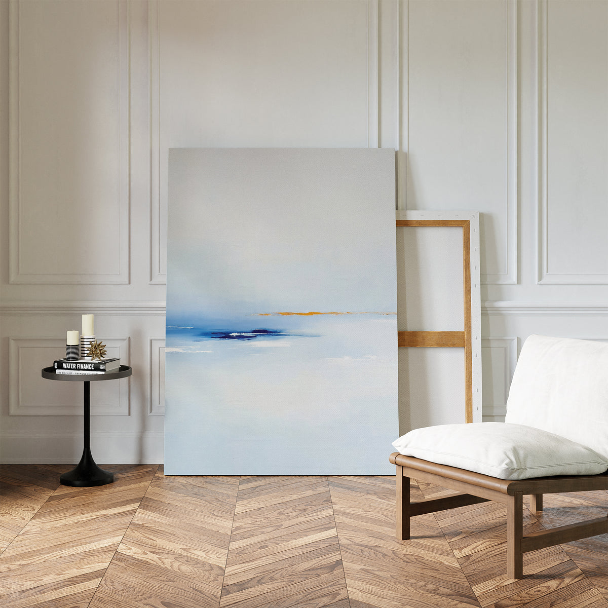 A minimalist living space with a white cushioned chair, black side table with candles and books, and the Distant Archipelago Canvas Art - Vertical in soft blue and white tones resting against a paneled wall on herringbone wood.