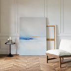 A minimalist living space with a white cushioned chair, black side table with candles and books, and the Distant Archipelago Canvas Art - Vertical in soft blue and white tones resting against a paneled wall on herringbone wood.