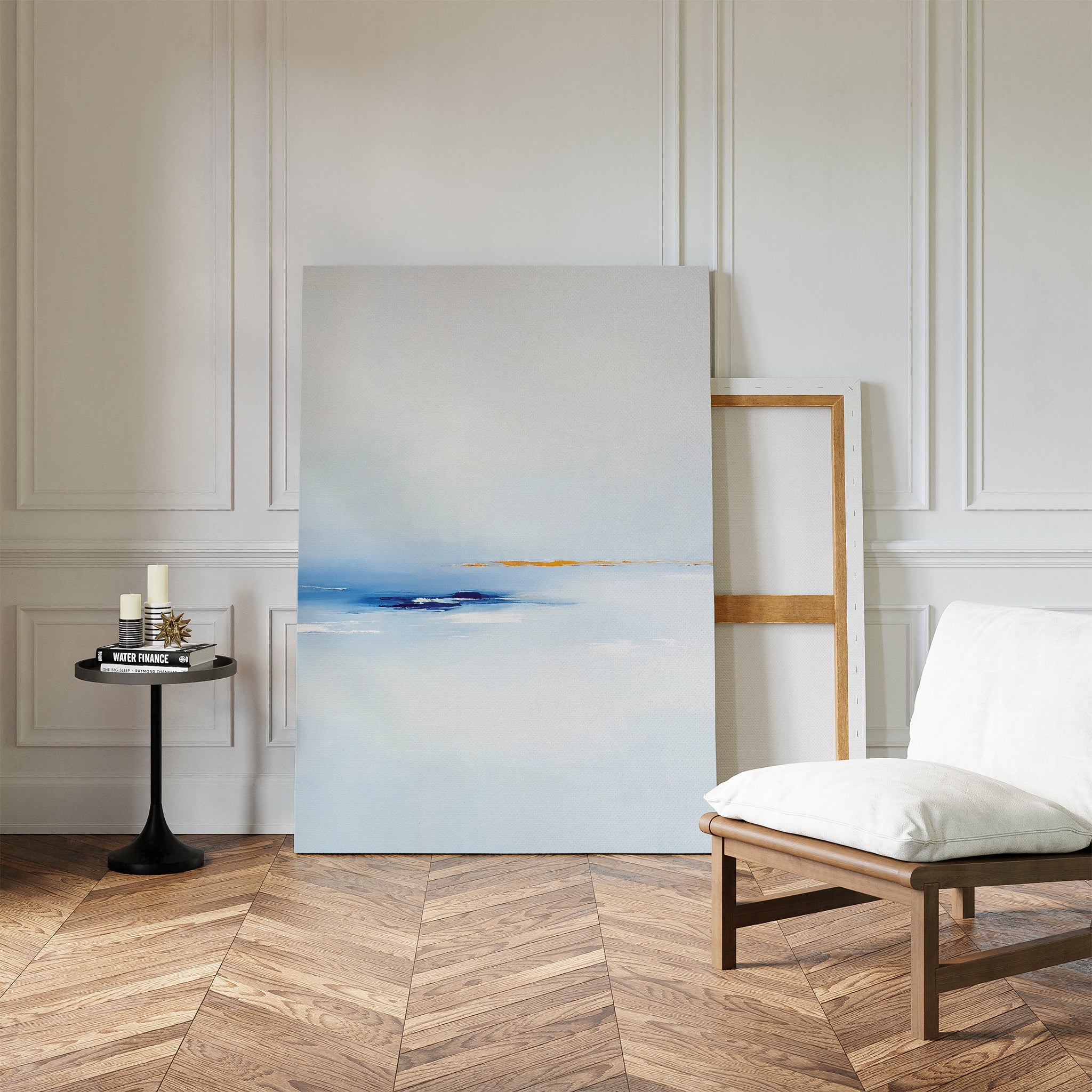 A minimalist living space with a white cushioned chair, black side table with candles and books, and the Distant Archipelago Canvas Art - Vertical in soft blue and white tones resting against a paneled wall on herringbone wood.