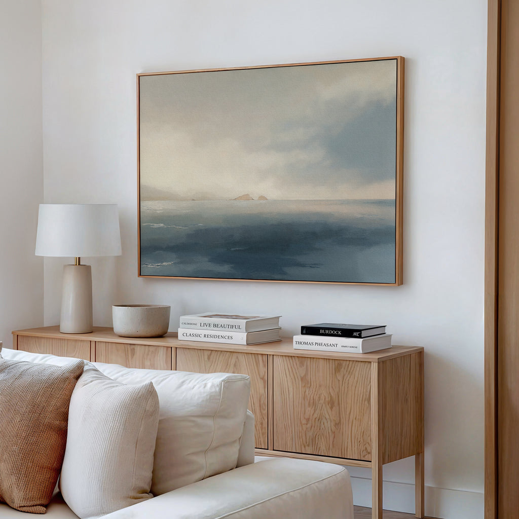 A minimal living room features a light wood sideboard with stacked books, a lamp, and a bowl. Above the beige sofa with pillows hangs the Distant Isles Canvas Art - Horizontal, creating tranquil home decor.