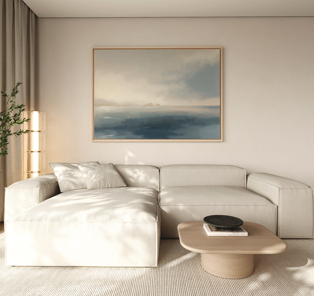 A modern living room with a light beige sectional, wooden coffee table, books, black bowl, tall plant, and the Distant Isles Canvas Art - Horizontal on a neutral wall creates a tranquil and serene home decor atmosphere.