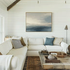 A bright coastal living room features a white sectional sofa, assorted pillows, a rustic wooden coffee table, and the Distant Isles Canvas Art - Horizontal on the white shiplap wall. Natural light streams in from a window.