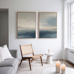 A modern minimalist living room with a light gray sofa, woven chair, round side table, and lit candles. The Distant Isles - Set of 2 serene seascape canvas prints adorn a white wall beside a tall window streaming in natural light.