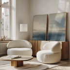 A modern living room features two rounded white chairs, a low wooden coffee table, a floor lamp, and the Distant Isles - Set of 2 seascape canvases leaning against a wood sideboard near a tall window.