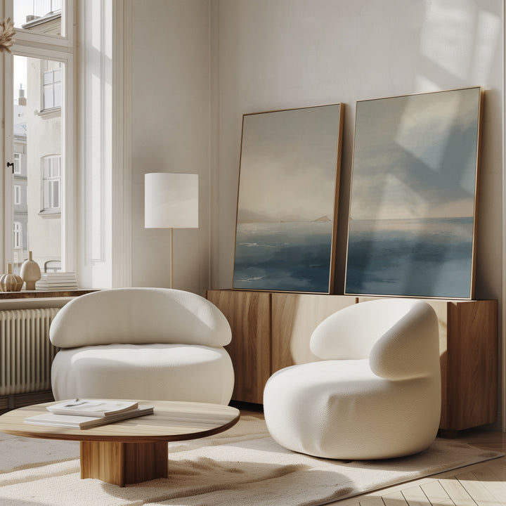 A modern living room features two rounded white chairs, a low wooden coffee table, a floor lamp, and the Distant Isles - Set of 2 seascape canvases leaning against a wood sideboard near a tall window.