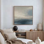 A minimalist living room with a beige sofa, neutral pillows, wooden sideboard, vases, books, and the Distant Isles Canvas Art - Square showcasing a serene seascape with distant mountains—Nordic Park style on a crisp white wall.