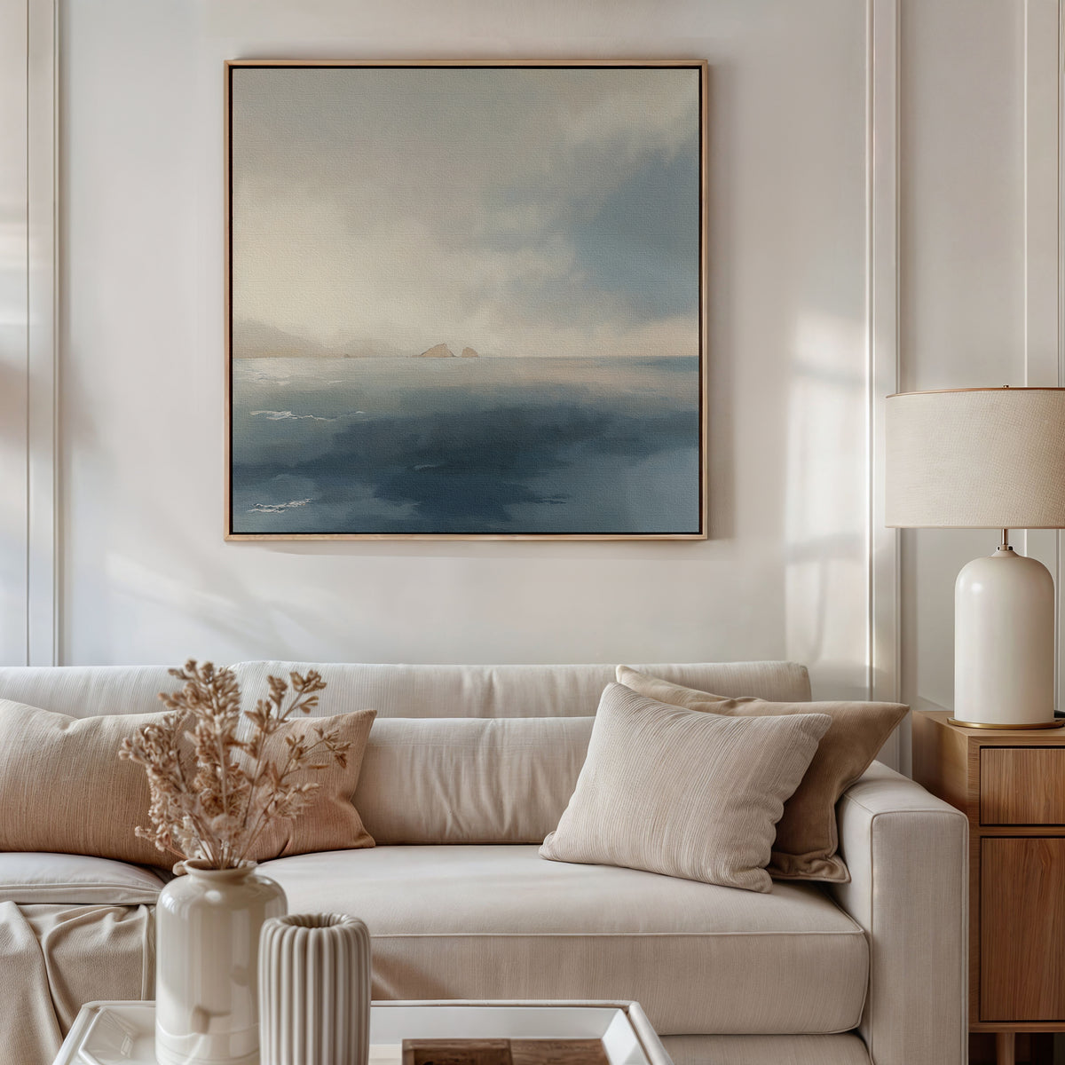 A cozy living room features a beige sofa with matching cushions, a wooden side table holding a white lamp and vase of dried flowers, while the Distant Isles Canvas Art - Square brings a calming touch above the sofa.