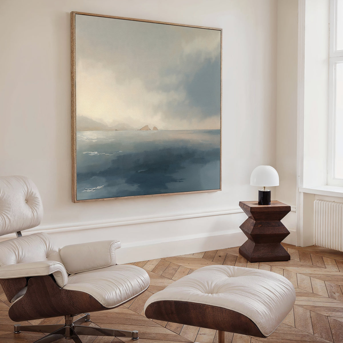 A cozy, modern living room features a cream lounge chair and ottoman, a wooden side table with a white lamp, and the Distant Isles Canvas Art - Square on the wall. Light streams in from a tall window, enhancing the tranquil ambiance.