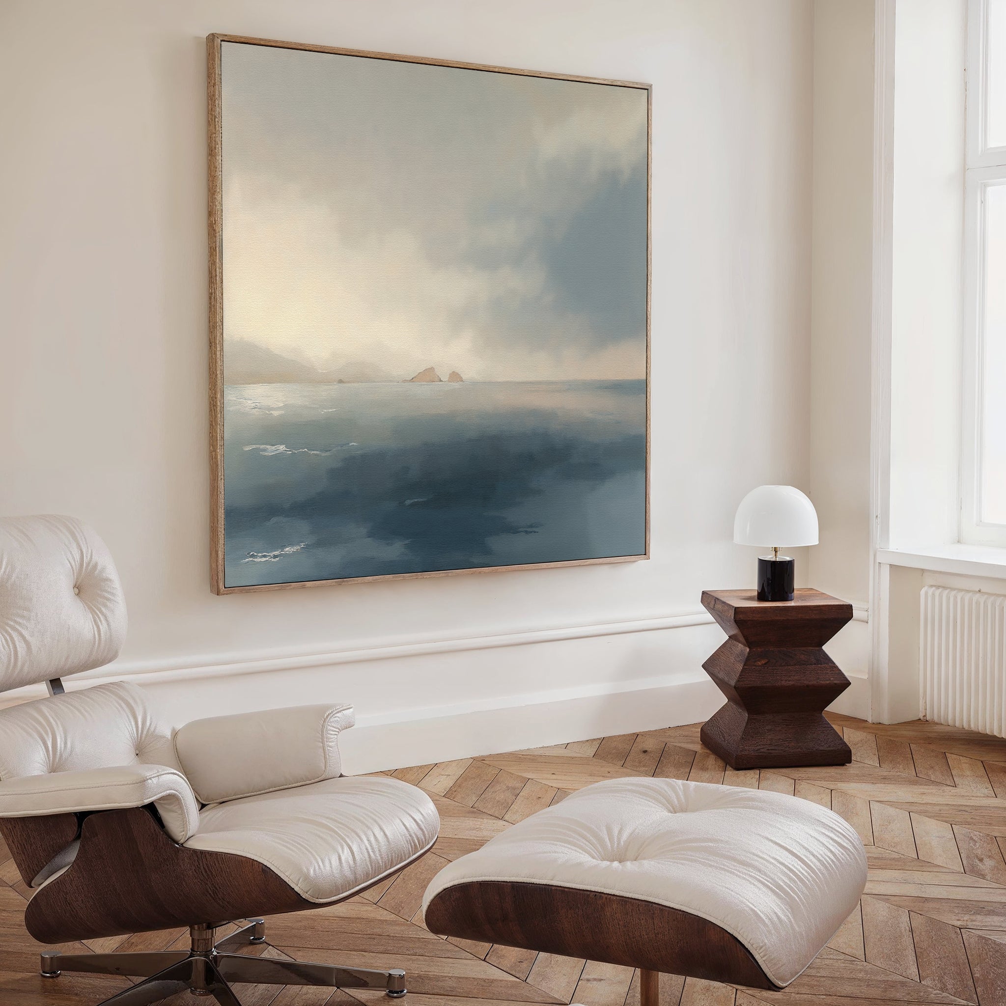 A cozy, modern living room features a cream lounge chair and ottoman, a wooden side table with a white lamp, and the Distant Isles Canvas Art - Square on the wall. Light streams in from a tall window, enhancing the tranquil ambiance.